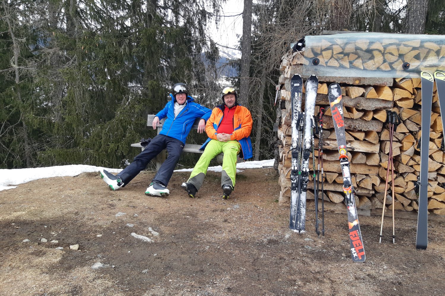 Zwei Skifahrer machen Pause auf einer Bank beim Holzstapel